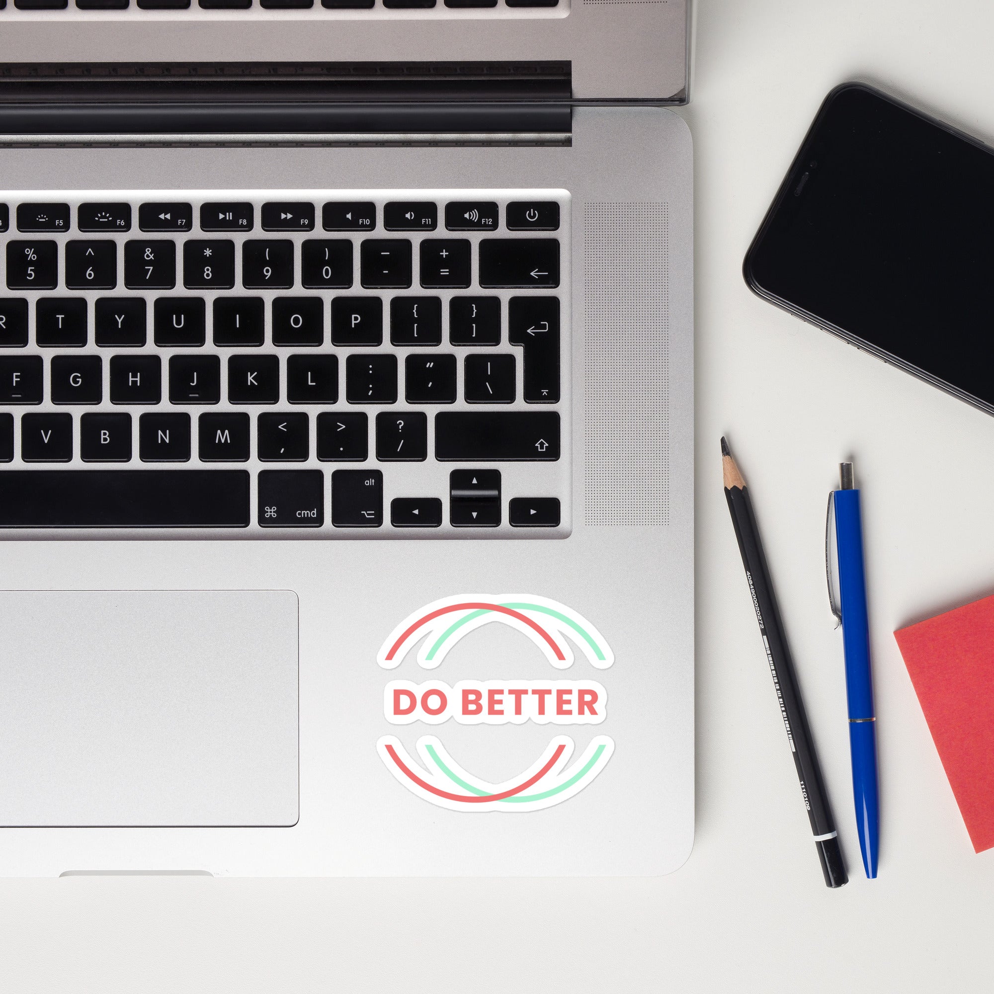 Scaling Better Do Better Sticker placed on silver laptop keyboard deck next to pencil and pen